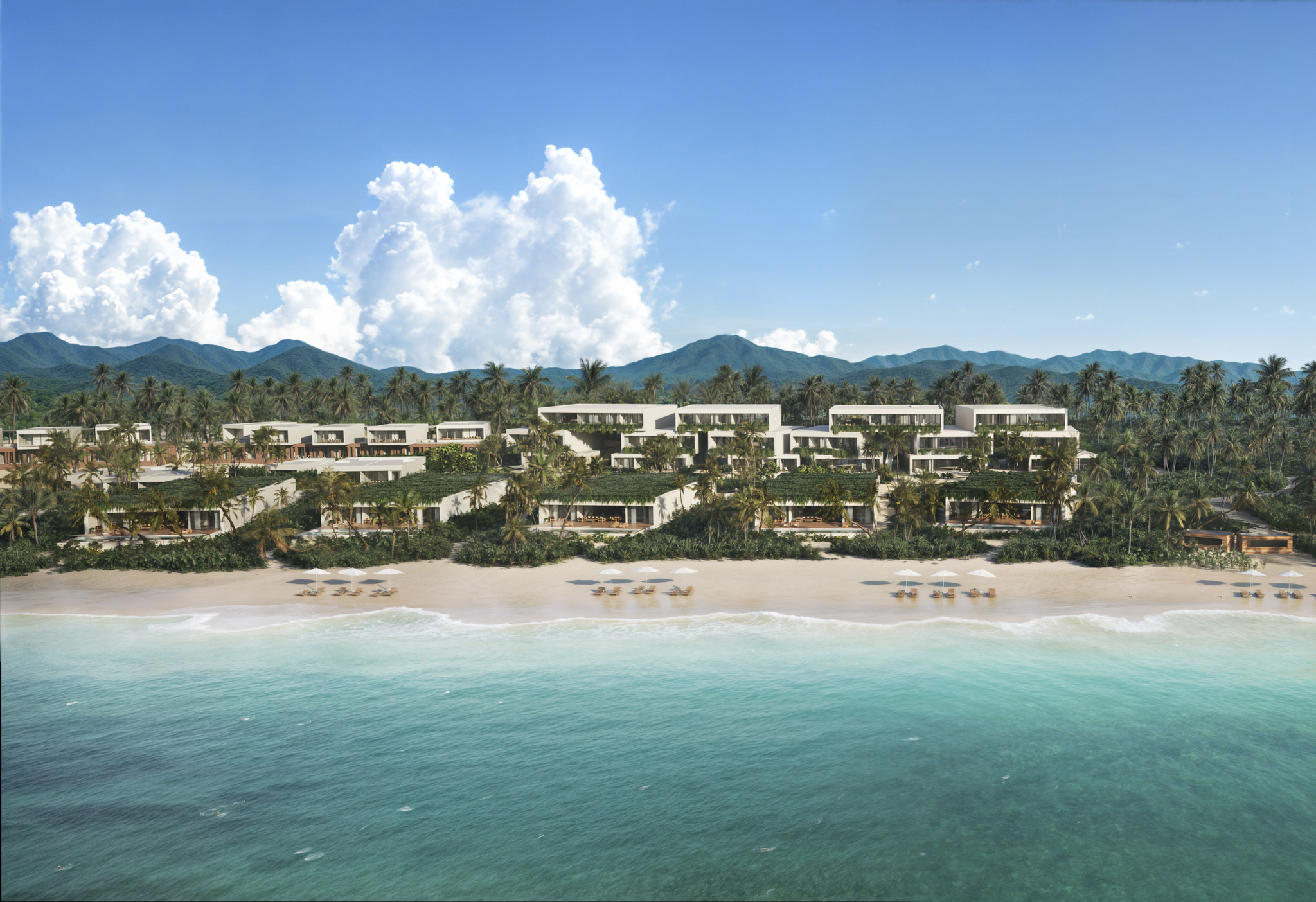 Aerial view of modern beachfront residences along a white-sand beach with turquoise water and lush hills under a bright blue sky.