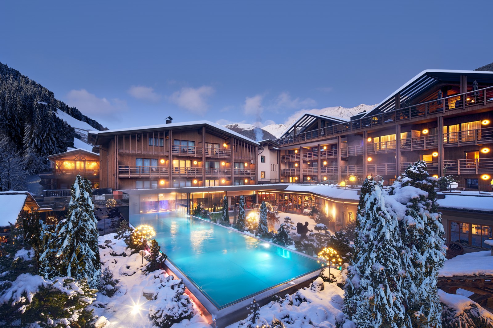 Snow-covered Alpine luxury resort with a heated outdoor pool glowing at dusk, surrounded by chalet-style buildings and illuminated balconies in the Dolomites.